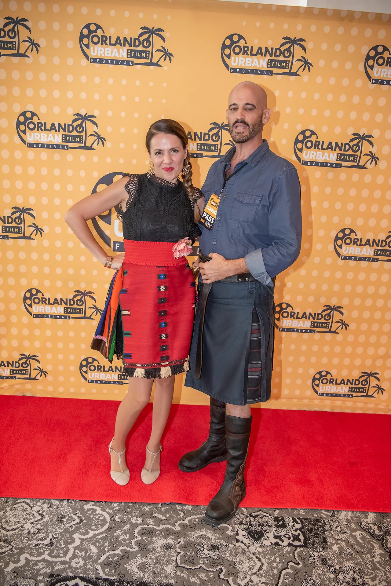 A couple posing on the red carpet, one in a red pleated skirt and one in a kilt