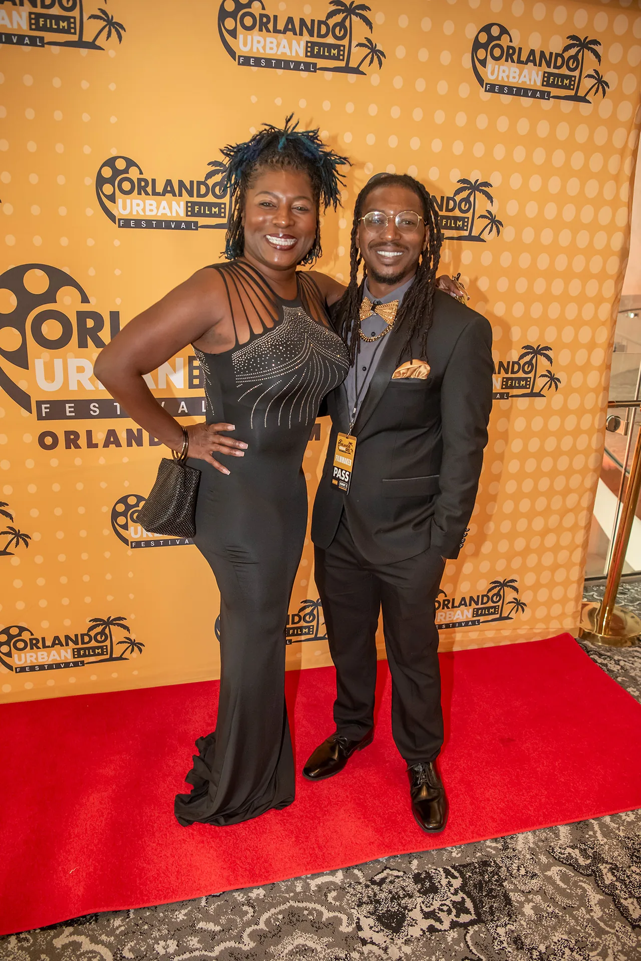 Two guests posing on the red carpet, one in a black gown with a feathered headpiece