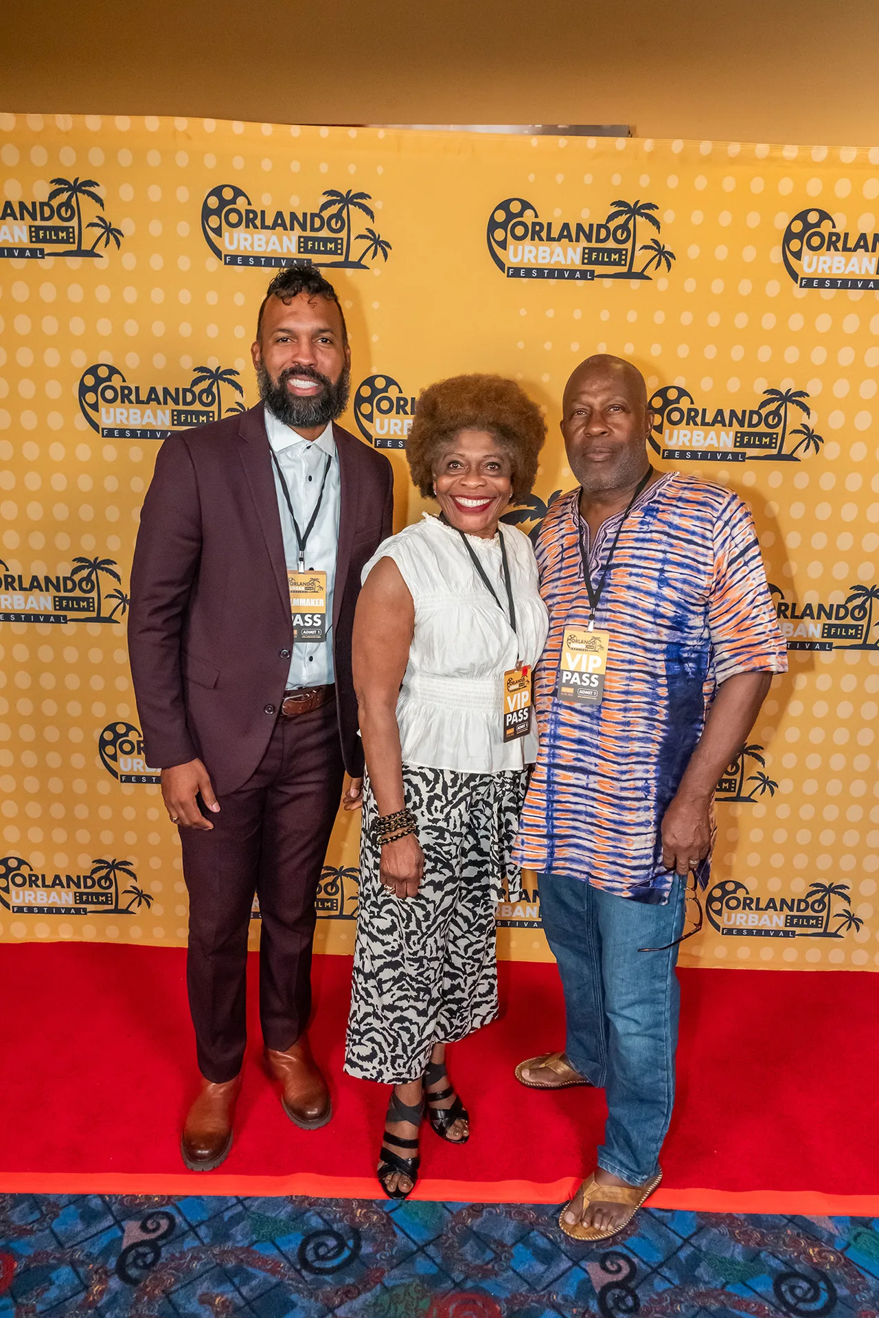 Three guests standing together on the red carpet, one in a maroon suit