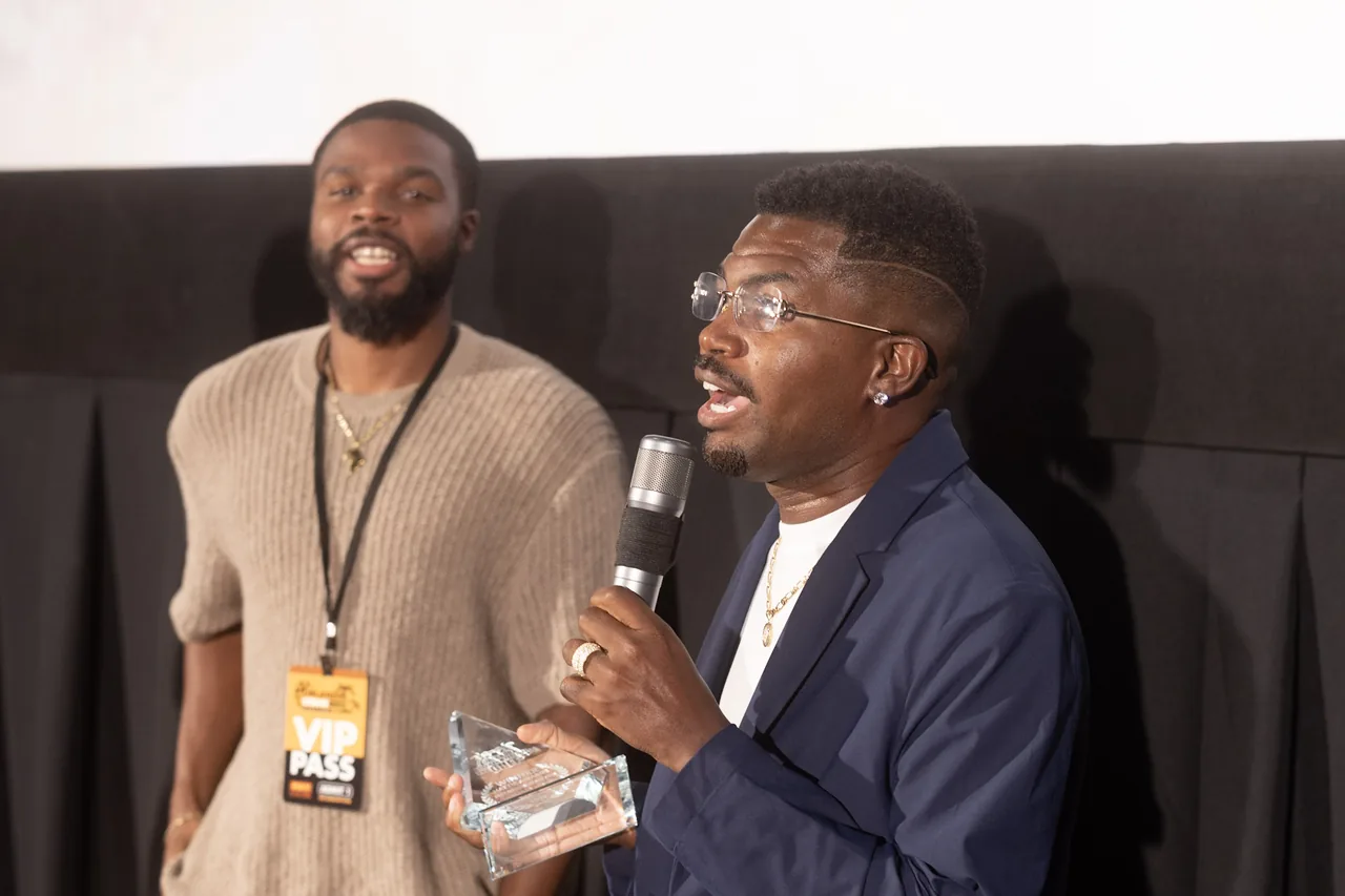 Two filmmakers on stage, one holding a crystal award, sharing a microphone